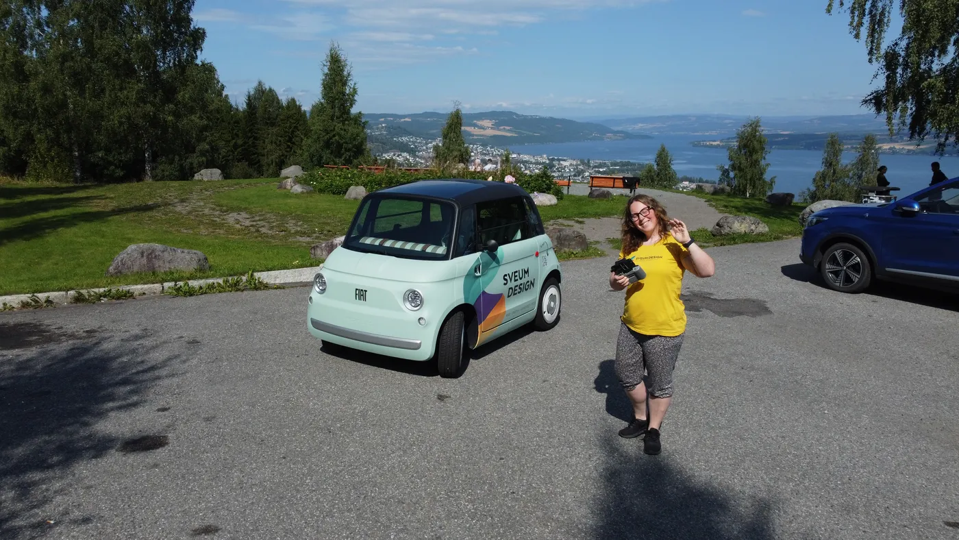 Kristine ved siden av den folierte Fiat Topolinoen med dronekontroller, ved utsikten over Mjøsa